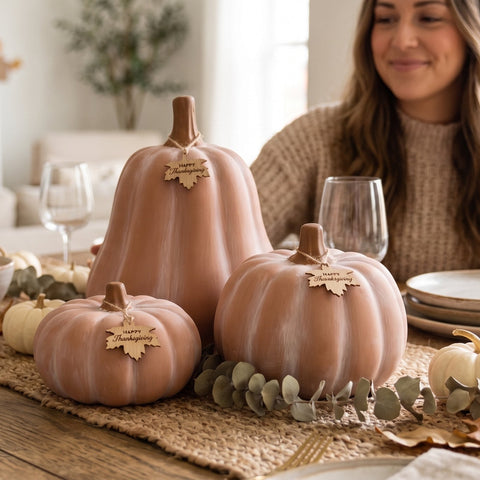 A woman in a cozy knit sweater sits at a wooden dining table decorated with three large blush pink pumpkins, smaller white pumpkins, a burlap runner, and fall leaves, in a bright, warm living room.